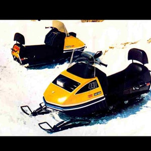 May include: Two yellow and black snowmobiles parked in the snow. The snowmobile in the foreground has a black seat and a 440cc engine. The snowmobile in the background has a yellow seat.