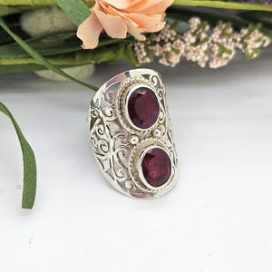 May include: A silver ring featuring two oval-shaped, deep red gemstones set vertically. The band is crafted with an intricate, openwork filigree design, and each stone is surrounded by a twisted rope-style metal border for a textured appearance.