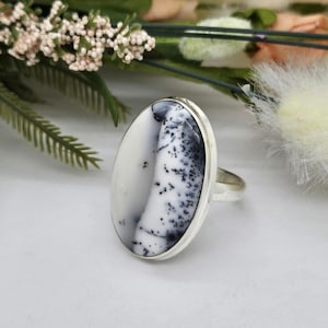 May include: A silver ring featuring a large, oval-shaped dendritic agate gemstone. The stone is primarily white with distinct, dark, fern-like inclusions that create a branching pattern across the surface. The metal band is smooth and polished.