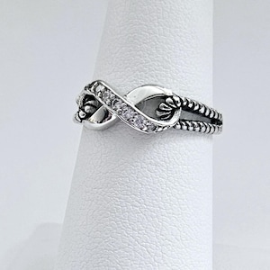 May include: A silver-toned ring in an infinity symbol design. The ring features a row of small, clear stones along one side of the infinity loop. The band has a twisted rope-like texture. The ring is displayed on a white surface.