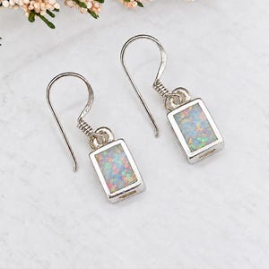 May include: A pair of silver drop earrings featuring rectangular settings with iridescent, multi-colored opal inlays. Each earring is suspended from a simple silver hook wire, showcasing a polished metal frame that surrounds the vibrant, shimmering stone.