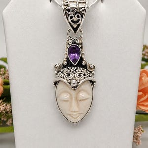 May include: A sterling silver pendant featuring a carved off-white face with closed eyes, topped with a teardrop-shaped purple amethyst gemstone and intricate scrollwork, suspended from a silver chain on a white display stand.