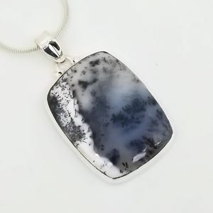 May include: A rectangular pendant with a dendritic agate stone, featuring a silver bezel and a silver chain. The stone displays a pattern of white, gray, and black, resembling a landscape. The pendant is a piece of jewelry.