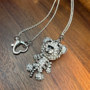 May include: A silver chain necklace with a rhinestone-covered teddy bear pendant. The bear has black eyes and a black outline.