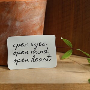 May include: A white card with the text "open eyes open mind open heart" written in black cursive. The card is sitting on a wooden surface next to a terracotta pot and a green plant.