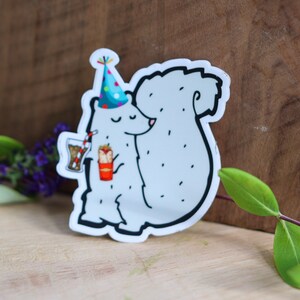 May include: A white sticker of a cartoon squirrel wearing a blue and white party hat, holding a red cup with a straw and a red and yellow food item. The squirrel is smiling and has a white tail.