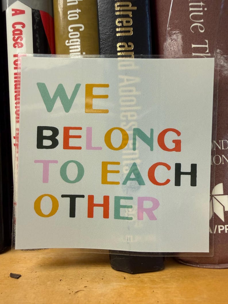 May include: A square sticker with the words "WE BELONG TO EACH OTHER" in various colors. The words are stacked vertically, with each word in a different color, including teal, yellow, black, red, orange, pink, and green.