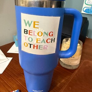 May include: A large blue Hydro Flask tumbler with a handle, lid, and straw. The tumbler features a white rectangle with the words "WE BELONG TO EACH OTHER" in colorful letters. The tumbler is on a wooden surface.