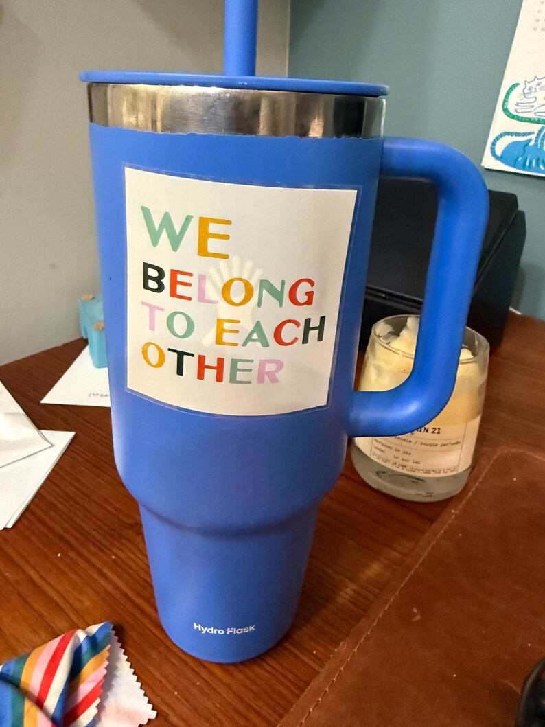 May include: A large blue Hydro Flask tumbler with a handle, lid, and straw. The tumbler features a white rectangle with the words "WE BELONG TO EACH OTHER" in colorful letters. The tumbler is on a wooden surface.