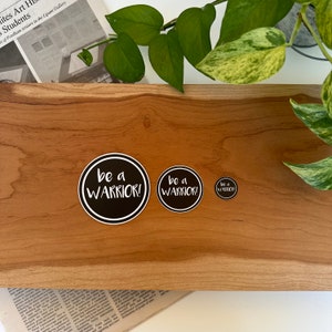 May include: Three black and white round stickers with the text "be a WARRIOR!" in white on a wooden surface. The stickers are different sizes.