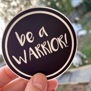 May include: A black and white round sticker with a white border. The sticker says "be a warrior!" in white text.