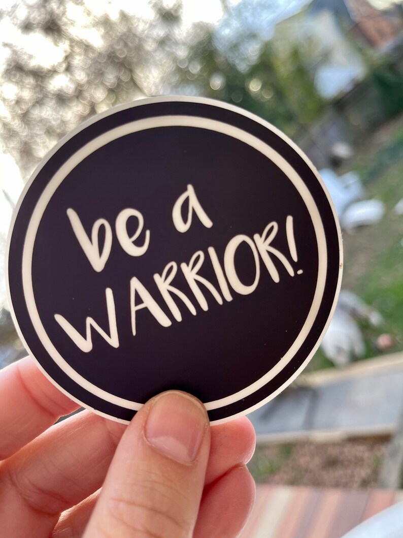 May include: A black and white round sticker with a white border. The sticker says "be a warrior!" in white text.