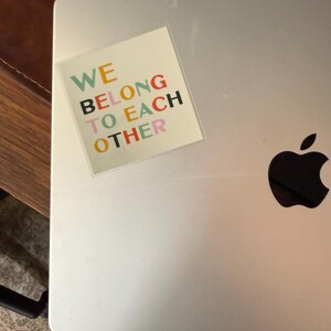 May include: A silver laptop with a colorful sticker that reads "WE BELONG TO EACH OTHER." The sticker has text in various colors, including green, orange, black, and pink. The Apple logo is visible on the laptop.