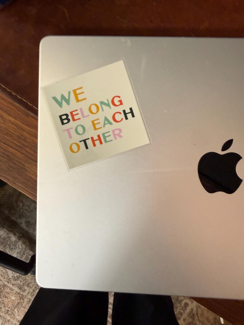 May include: A silver laptop with a colorful sticker that reads "WE BELONG TO EACH OTHER." The sticker has text in various colors, including green, orange, black, and pink. The Apple logo is visible on the laptop.