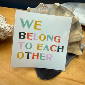 May include: A square sticker with the words "WE BELONG TO EACH OTHER" in various colors: teal, yellow, black, red, orange, and pink. The sticker is on a wooden surface, with a seashell in the background.