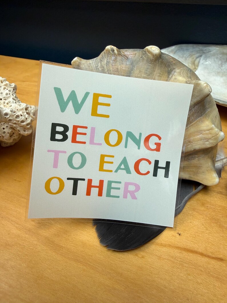 May include: A square sticker with the words "WE BELONG TO EACH OTHER" in various colors: teal, yellow, black, red, orange, and pink. The sticker is on a wooden surface, with a seashell in the background.