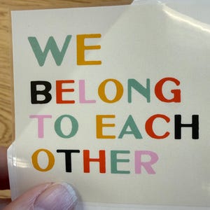 May include: A sticker with the words "WE BELONG TO EACH OTHER" in various colors. The text is in a sans-serif font and is arranged in four lines. The sticker is on a white background.