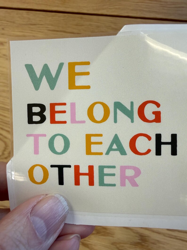 May include: A sticker with the words "WE BELONG TO EACH OTHER" in various colors. The text is in a sans-serif font and is arranged in four lines. The sticker is on a white background.