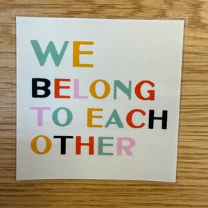 May include: A square sticker with the words "WE BELONG TO EACH OTHER" in various colors. The text is arranged in three lines, with each word in a different color, including teal, yellow, black, orange, pink, and red.