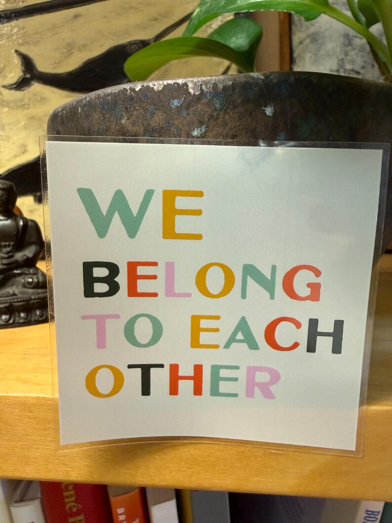 May include: A white card with the words "WE BELONG TO EACH OTHER" in colorful block letters. The card is in a clear plastic sleeve and is displayed on a wooden shelf. A dark brown pot with a plant is in the background.