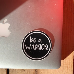 May include: A black and white round sticker with the text "be a warrior!" in white lettering on a black background.