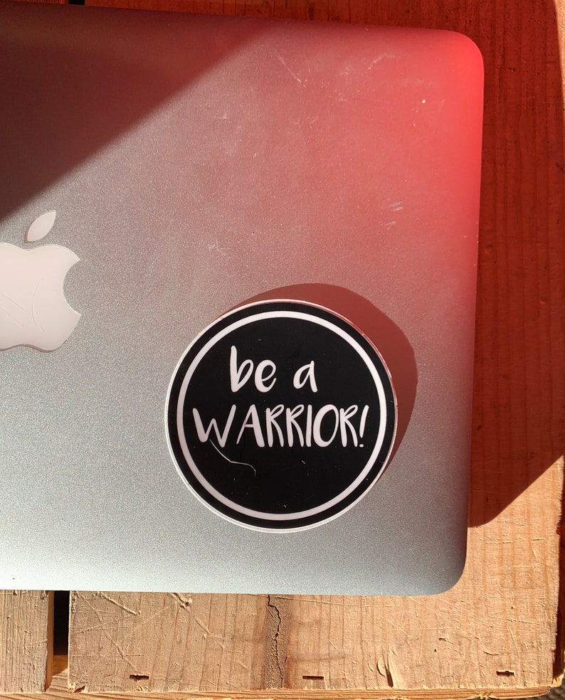 May include: A black and white round sticker with the text "be a warrior!" in white lettering on a black background.