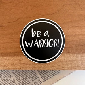May include: A black and white round sticker with a white border. The sticker has the text "be a WARRIOR!" in white lettering on a black background.