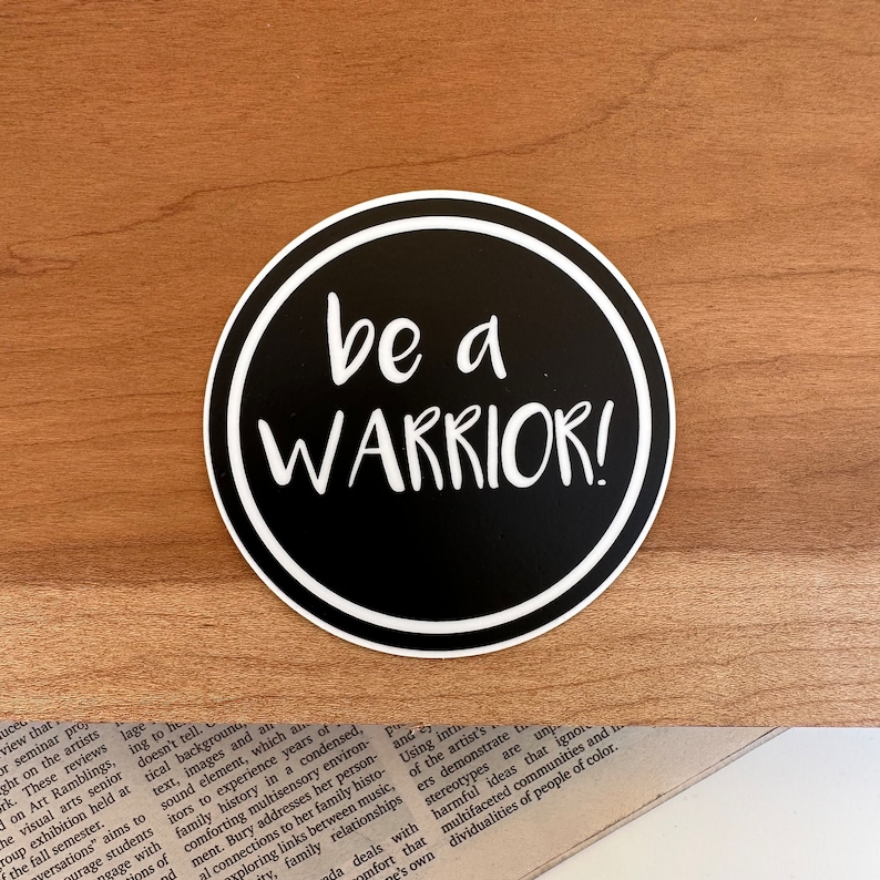 May include: A black and white round sticker with a white border. The sticker has the text "be a WARRIOR!" in white lettering on a black background.