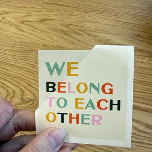 May include: A square sticker with the words "WE BELONG TO EACH OTHER" in various colors. The text is in a bold, sans-serif font. The sticker is held by a hand, and the background is a wooden surface.