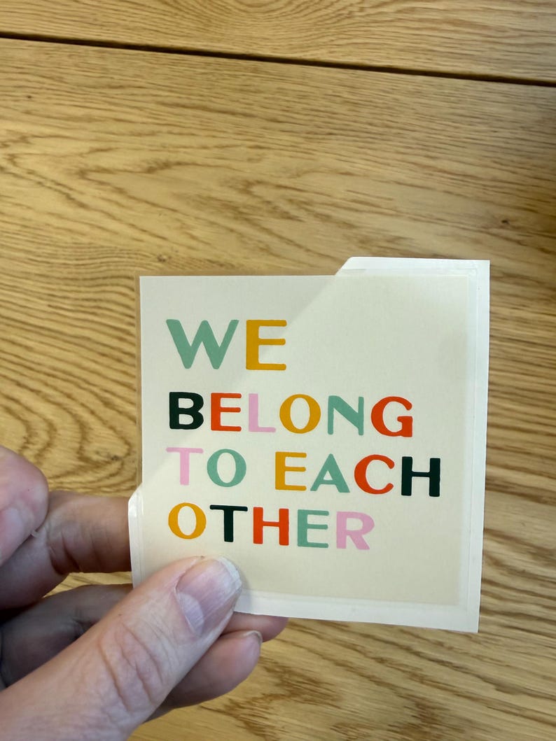 May include: A square sticker with the words "WE BELONG TO EACH OTHER" in various colors. The text is in a bold, sans-serif font. The sticker is held by a hand, and the background is a wooden surface.