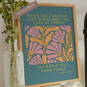 Op de afbeelding: Een ingelijste kunstdruk met een teal achtergrond en een bloemenontwerp in roze en oranje. De print bevat de quote "Wanneer de kracht van liefde de liefde voor macht overwint, zal de wereld vrede kennen" van Jimi Hendrix. De lijst is van lichtbruin hout.