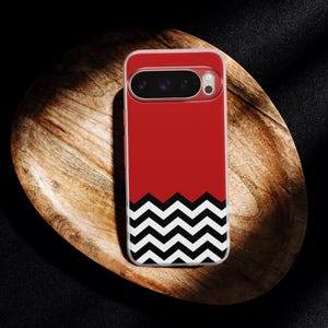 Può includere: Custodia per telefono rossa con un motivo a zig-zag bianco e nero nella parte inferiore. La custodia ha un bordo trasparente e un ritaglio per la fotocamera. La custodia è su un piatto di legno.