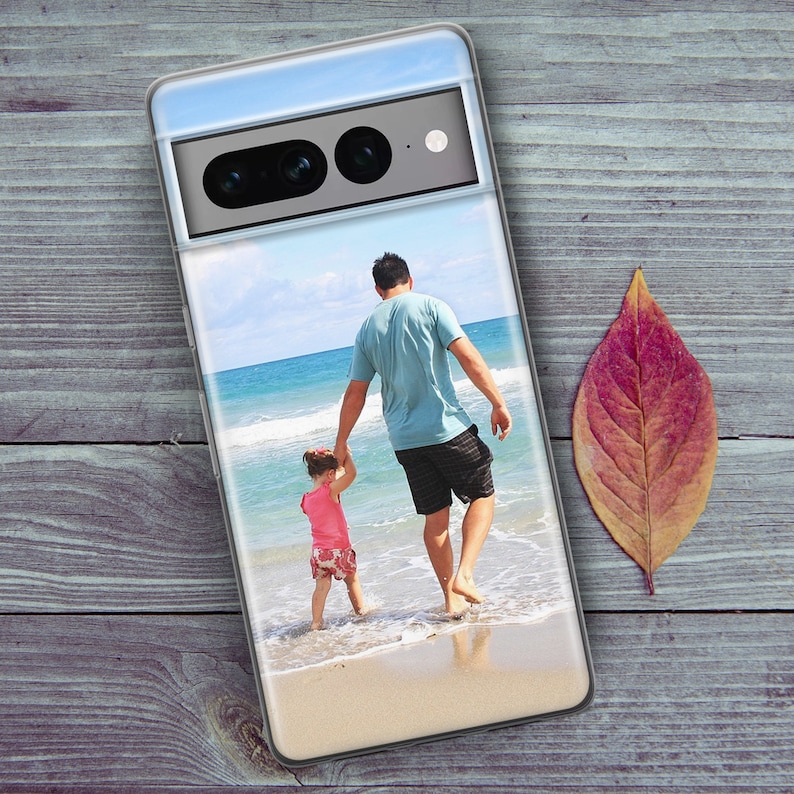 Może przedstawiać: Przezroczyste etui na telefon z fotografią ojca i c&oacute;rki spacerujących po plaży. Zdjęcie przedstawia błękitne niebo, białe chmury i błękitną wodę oceanu. Ojciec ma na sobie niebieską koszulę i czarne spodenki. C&oacute;rka ma na sobie r&oacute;żową koszulę i białe spodenki.