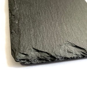 Puede incluir: Una tabla de servir de pizarra gris oscuro con una superficie rugosa y texturizada. La tabla tiene una forma natural irregular con un borde ligeramente curvo.