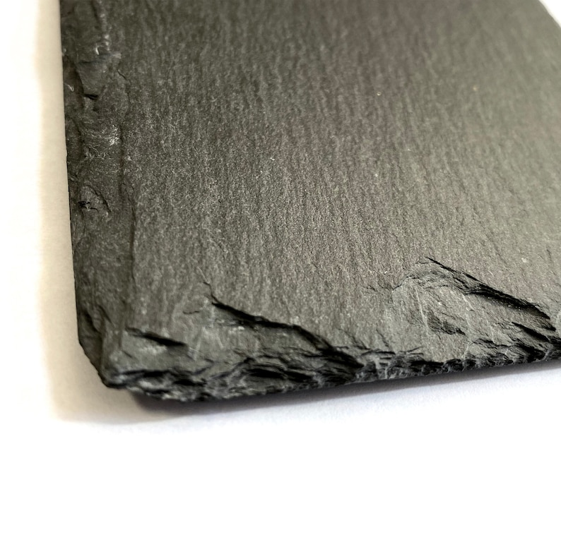 Puede incluir: Una tabla de servir de pizarra gris oscuro con una superficie rugosa y texturizada. La tabla tiene una forma natural irregular con un borde ligeramente curvo.