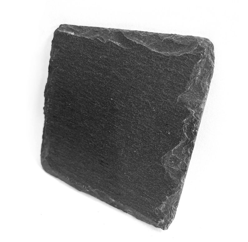 Puede incluir: Una tabla de servir de pizarra negra con una superficie rugosa y texturizada. La tabla es rectangular y tiene bordes ligeramente irregulares.