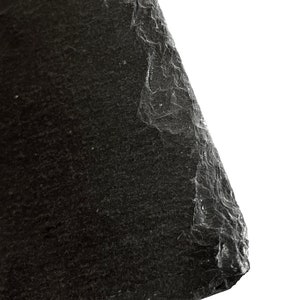 Puede incluir: Primer plano de una roca de pizarra gris oscuro con una superficie rugosa y texturizada. La roca tiene un borde irregular y est&aacute; inclinada hacia el lado derecho de la imagen.