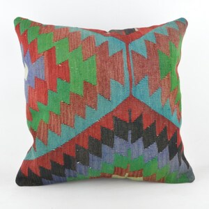 Puede incluir: Cojín cuadrado con un patrón geométrico en rojo, verde, azul y negro. El cojín tejido tiene un diseño simétrico, ideal para la decoración del hogar.
