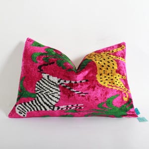 Puede incluir: Un cojín decorativo con un fondo rosa vibrante. El cojín presenta un diseño con una cebra, un leopardo y follaje verde. El leopardo tiene manchas amarillas. El cojín está hecho de un material suave y aterciopelado.