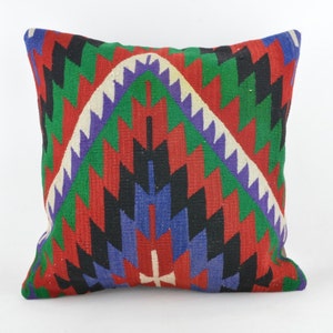 Puede incluir: Cojín cuadrado con un patrón geométrico en rojo, verde, negro, azul, morado y blanco. El cojín presenta un diseño central de un arco estilizado con un borde en zigzag. El cojín es probablemente de tela tejida.