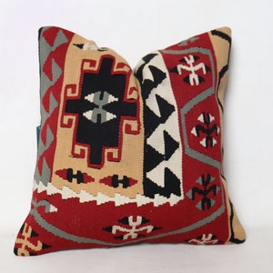 Puede incluir: Cojín cuadrado decorativo con un patrón geométrico en rojo, negro, beige, gris y blanco. El cojín presenta un diseño tejido con varias formas y símbolos, adecuado para la decoración del hogar.