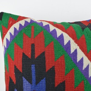 Puede incluir: Primer plano de un cojín decorativo con un vibrante patrón geométrico. El diseño presenta zigzags en rojo, verde, morado, negro y azul, sobre un fondo blanco. El cojín es de forma cuadrada.