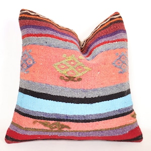 Puede incluir: Un cojín cuadrado tejido con un patrón de rayas vibrantes. El cojín presenta bandas de rojo, naranja, morado, gris, negro y azul. El centro tiene un campo de color coral con diseños geométricos bordados en verde y marrón.