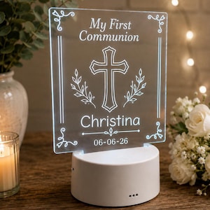 May include: A clear acrylic night light with the text "My First Communion", a cross, and the name "Christina" with the date 06-06-26. The light sits on a white base and is surrounded by decorative elements.