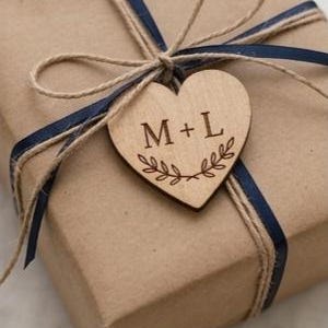 May include: A square gift box wrapped in brown paper, tied with a dark blue ribbon and natural twine. A wooden heart-shaped tag is attached to the center, featuring the engraved text M + L above a decorative leaf branch illustration.