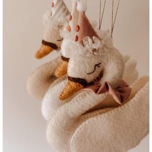May include: Close-up of two white felt swan ornaments with party hats. The hats are striped and dotted with small orange circles. Each swan has a brown beak, a pink bow, and a white lace collar. The ornaments are suspended by thin gold strings.
