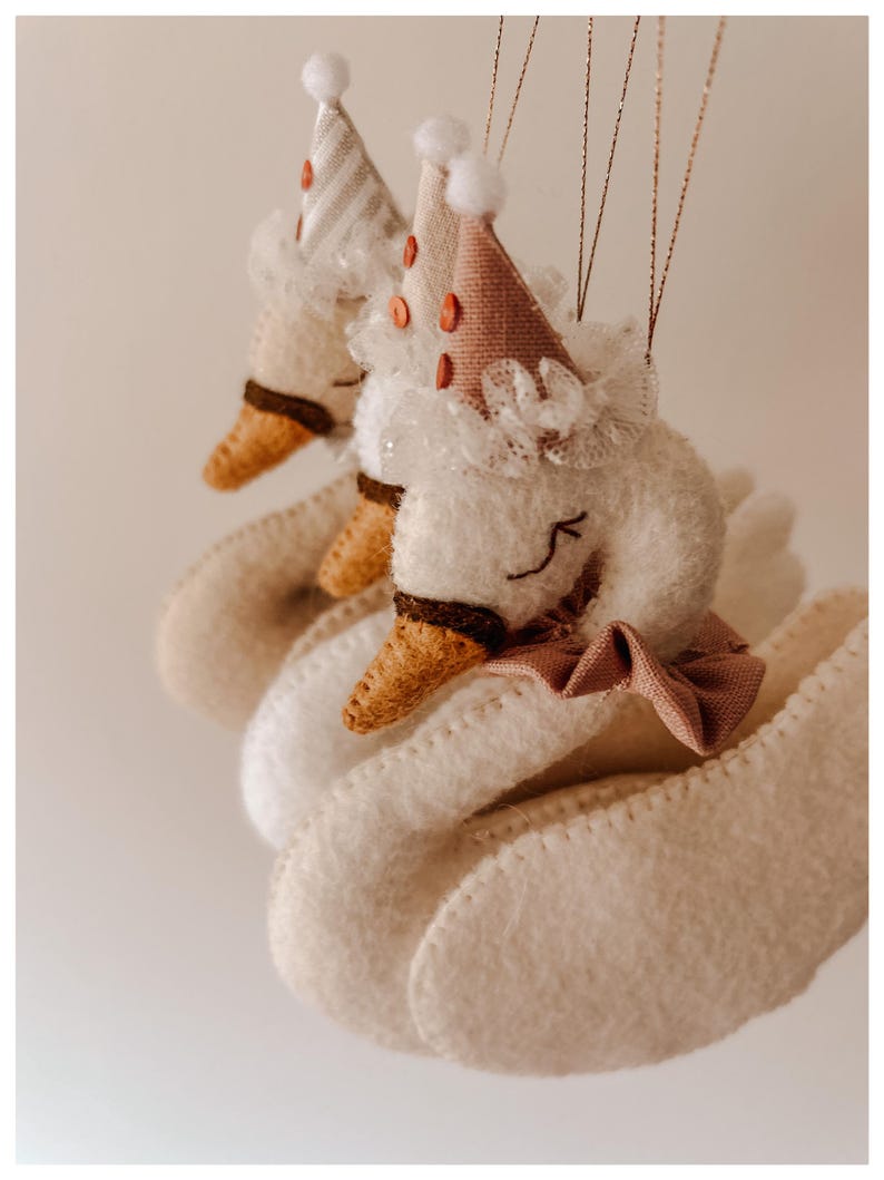 May include: Close-up of two white felt swan ornaments with party hats. The hats are striped and dotted with small orange circles. Each swan has a brown beak, a pink bow, and a white lace collar. The ornaments are suspended by thin gold strings.