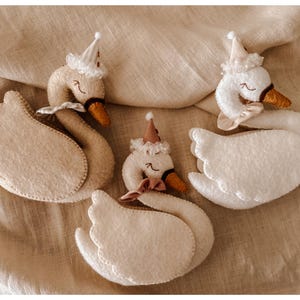 May include: Four handmade felt swan ornaments. Each swan has a white body, beige wings, and a brown beak. They are adorned with small party hats and bows. The swans are arranged on a beige fabric background.