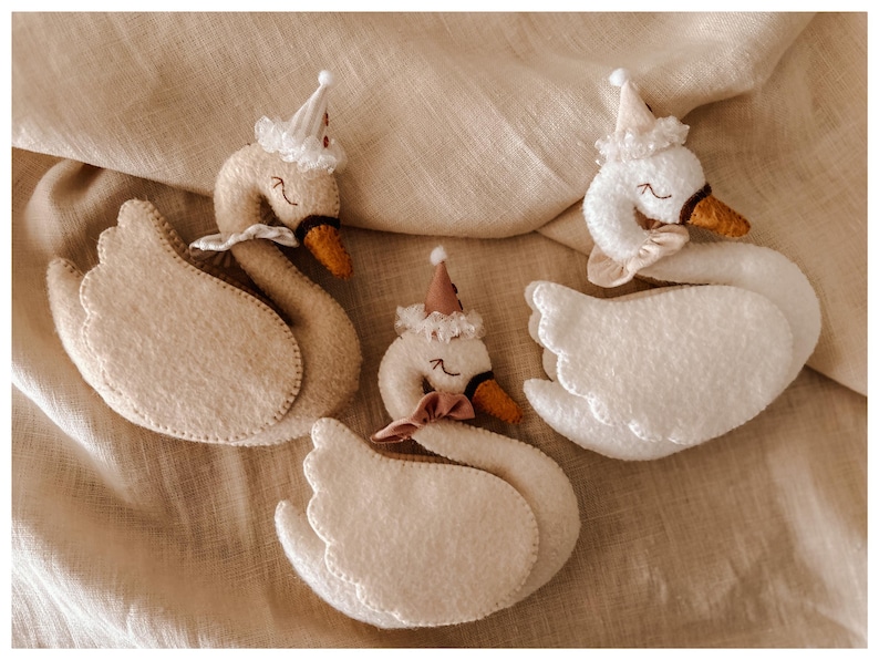 May include: Four handmade felt swan ornaments. Each swan has a white body, beige wings, and a brown beak. They are adorned with small party hats and bows. The swans are arranged on a beige fabric background.