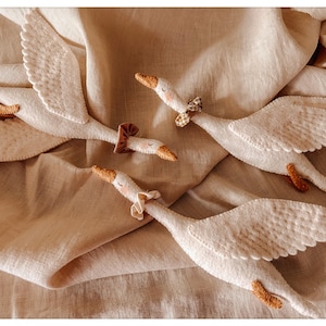 May include: Four white felt goose toys with outstretched wings and small, decorative bows around their necks. The geese have orange beaks and feet, and are arranged on a beige fabric background.